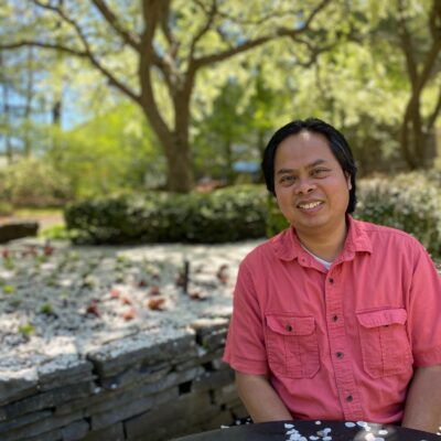 A brown-skinned Cambodian man wearing a bright pink short-sleeve shirt sits in a park, smiling at the camera. A large tree fills the background.