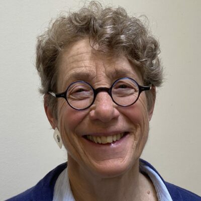 Poet Rebekah Wolman, a white Jewish woman with short curly hair and round-rimmed glasses, smiles widely at the camera