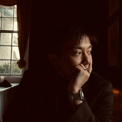 A person in a black blazer sits in a soft window light, their chin resting on their left hand while gazing out the window.