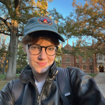 A white, queer person, wearing glasses, a blue hat, a black leather jacket, and a backpack. They are smiling at the camera, with autumnal trees and a blue sky in the background.