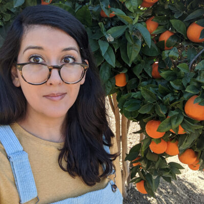 The poet Laura Villareal wears large glasses, light denim overalls, and a light mustard-colored shirt. She has long wavy black hair and stands in front of an orange tree.