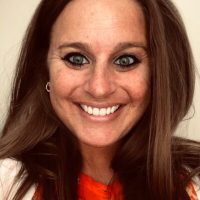 A close-up portrait of the author, a white woman, smiling into the camera against a beige backdrop. She has blue eyes, long brown hair, and is wearing an orange floral print blouse.