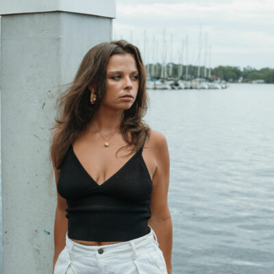 A Caucasian woman stands by a waterfront, looking to the side. She wears a sleeveless v-neck black top, gold pendant, and dangling earrings. She has wavy brown hair that falls past her shoulders.