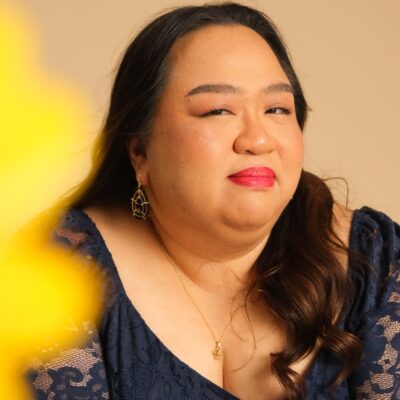 The poet, a Filipino woman with long dark hair, looks at the camera with a slight smile. She is wearing a black dress and a small pendant on a gold chain.