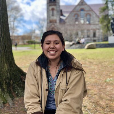 A young Diné woman wearing a khaki jacket sits underneath a tree outside on a college campus, smiling widely. She has long straight dark hair and is wearing a grey v-neck shirt and a double-strand turquoise necklace. There is a stone building in the distance and the leaves on the ground are brown.