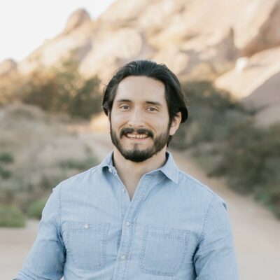 A dark-haired Latinx man with a beard and a chambray button-down shirt smiles as he stands outdoors.