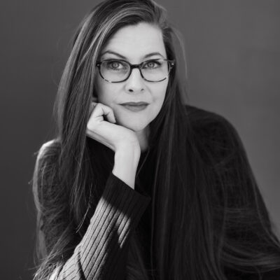 A black-and-white photograph of a Caucasian woman with long hair parted at the side. She wears glasses and is propping her chin in her right hand while looking at the camera.
