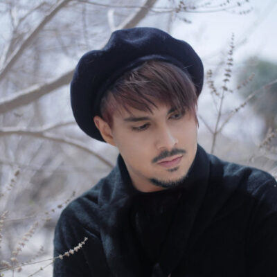 A person with short, reddish-brown hair & a neatly trimmed goatee wears a black beret & a black sweater, standing among leafless branches.