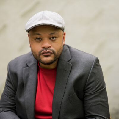 A photo of Christian J. Collier, a Black Poet, wearing a red t-shirt, grey blazer, and off-white flat cap. He is looking directly into the camera.