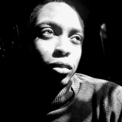 A black-and-white photo of a Black man gazing up to the right. His face is bathed in light, his gaze is soft, and he wears a turtleneck sweater.