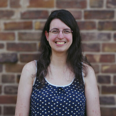 A fair-skinned woman with wavy dark brown hair that falls past her shoulders stands against a brick wall, smiling. She wears glasses and a scoop-neck sleeveless navy top patterned with small white hearts.