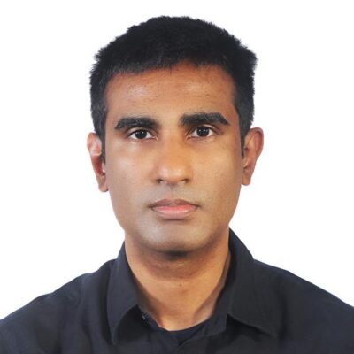 A Sri Lankan man looks at the camera with an even expression. He has short cropped black hair and thick eyebrows, and wears a black collared shirt.