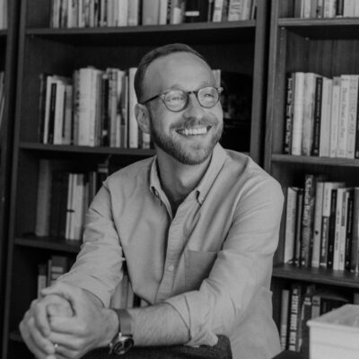 The poet Bobby Elliott, a Caucasian man with close-cropped light brown hair and a light brown heard, looks off to the side, smiling. His hands are clasped together in a relaxed pose and he wears brown round-rimmed glasses, a wristwatch, and a light blue button-down shirt. He sits in front of two large shelves of books.