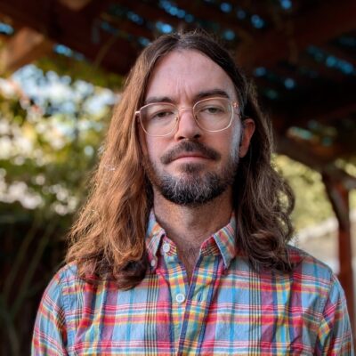 Nicholas Yingling stands outdoors under a wooden structure, looking at the camera with a neutral expression on his face. He wears wire-rimmed glasses and a colorful button-down checkered shirt. He has a beard, thick dark brown eyebrows, and wavy brown hair that falls to his shoulders.